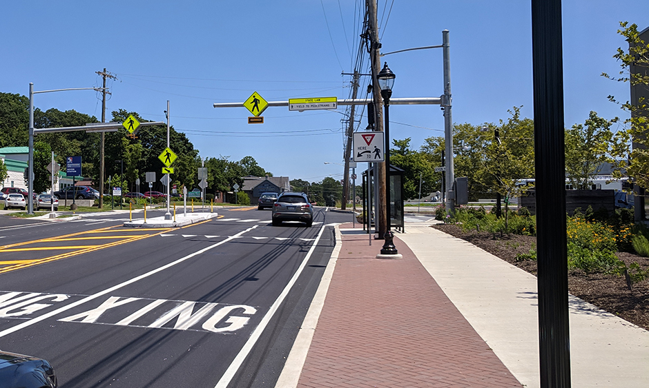 A sidewalk alongside a road with clearly painted crosswalk markers and a street sign indicating a pedestrian crossing
