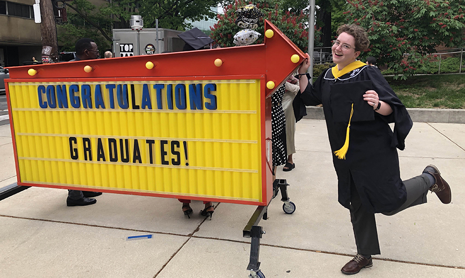DVRPC staff member Kaylen Phillips in a graduation gown standing next to a sign that reads, "Congratulations graduates!"