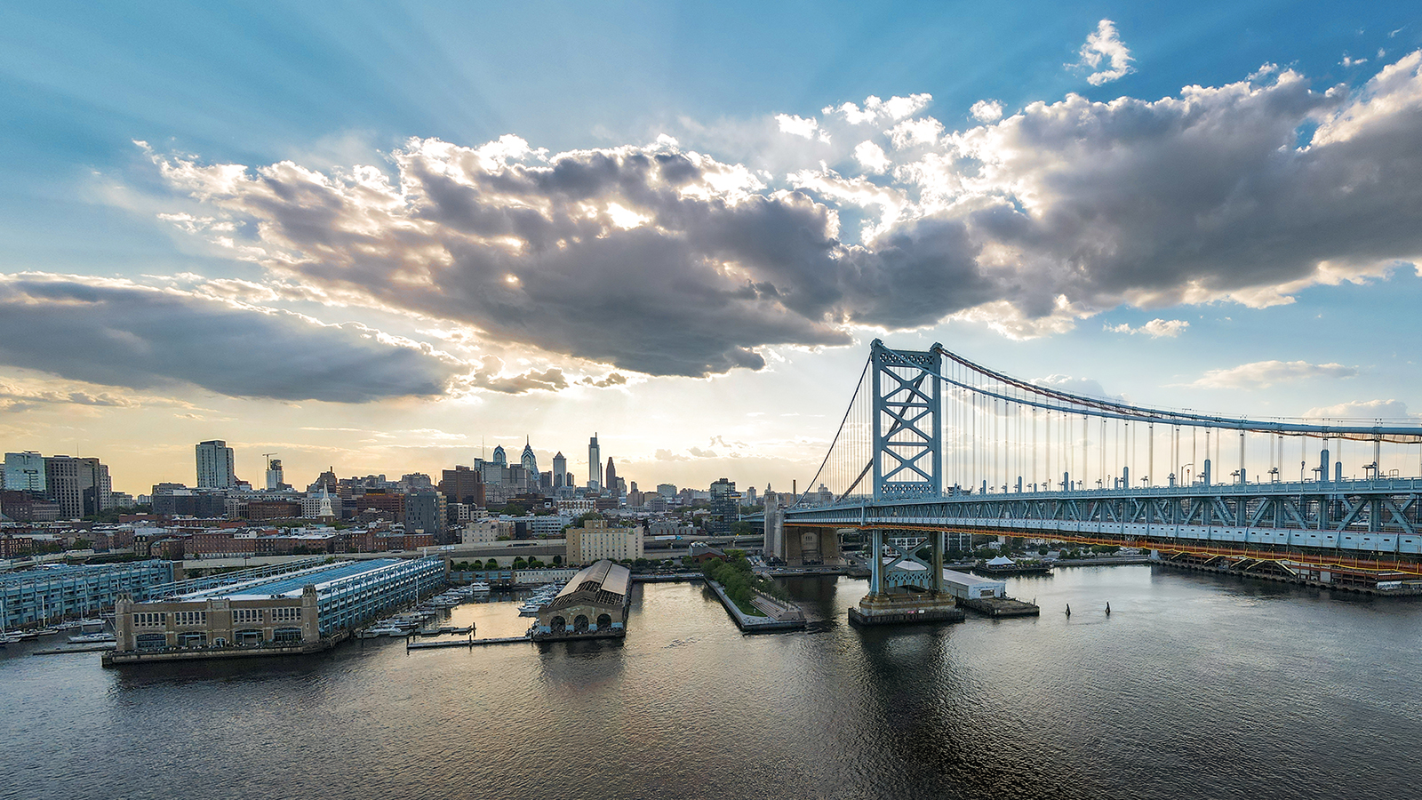 Big white clouds appear over the City of Philadelphia skyline. The Delaware River and Ben Franklin Bridge are in the foreground.