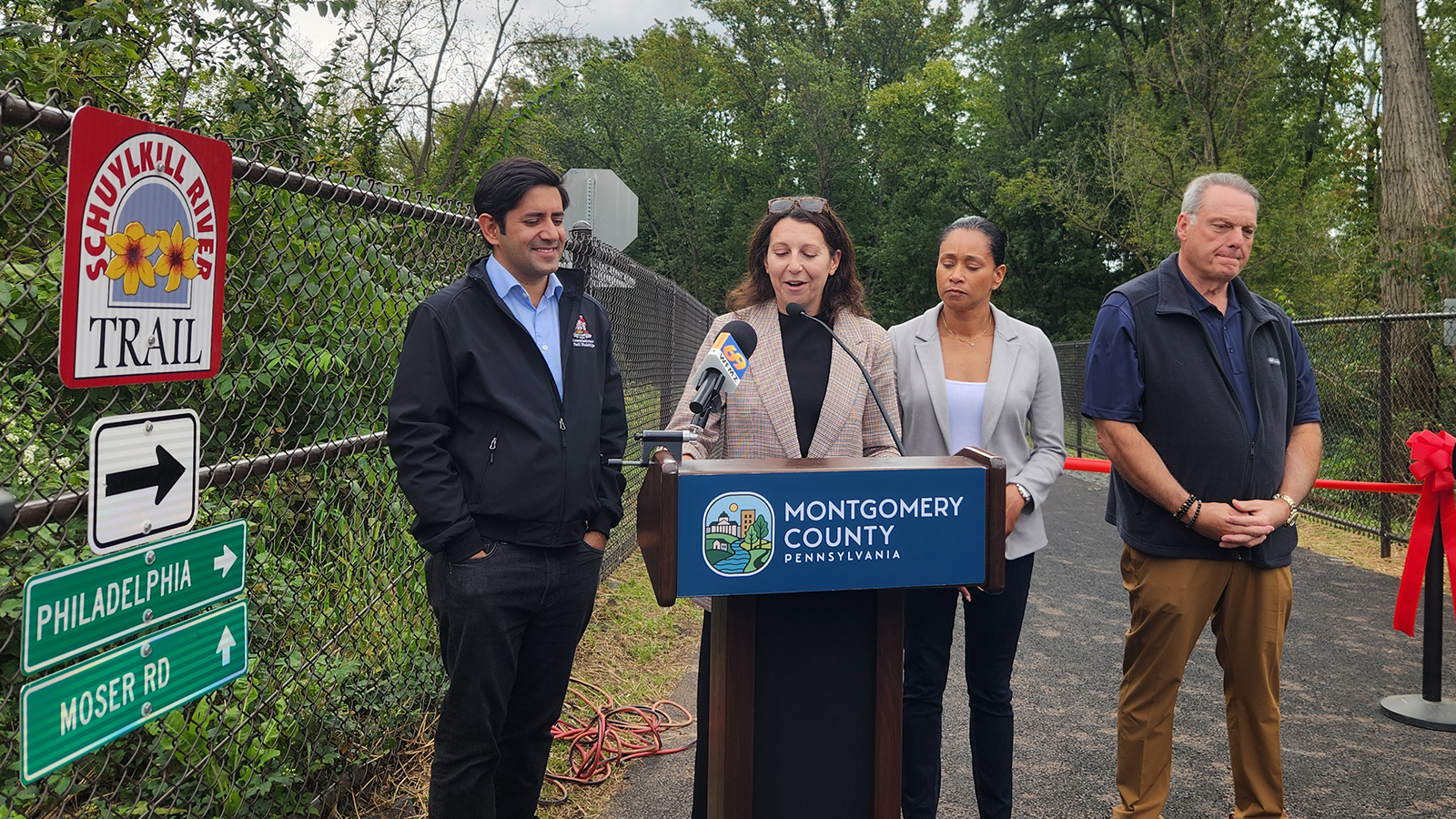 DVRPC Executive Director Ariella Maron speaks at a podium outdoors during a trail opening ceremony. She is surrounded by other officials.