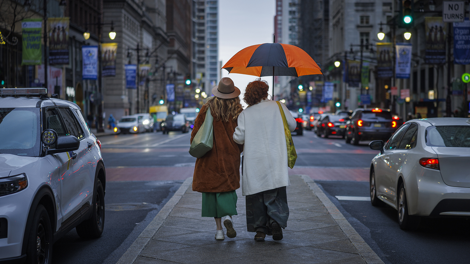Two female pedestrians walk along the median on Broad Street in Center City, Philadelphia, on an overcast day. One woman holds an umbrella, and traffic passes the pedestrians on either side of the median.